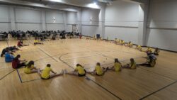 Group of girls in a gym, sitting in a straddle stretch, in a circle.