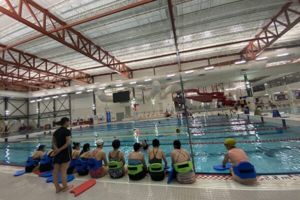 Backs of women in swimming suits sitting on the edge of a pool deck listening to a swim instructor who is in the water.
