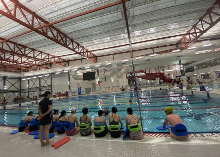 Backs of women in swimming suits sitting on the edge of a pool deck listening to a swim instructor who is in the water.