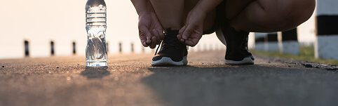 A person from the knees down, bent down and tying their black running shoe, on the pavement.