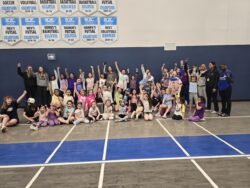 Group of 50 girls in a gym posing for a picture in athletic wear and their arms raised with a fist pump