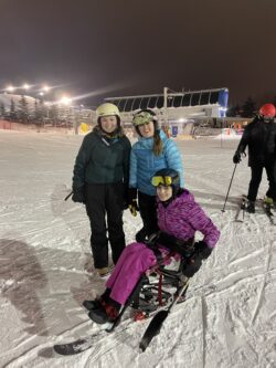 One women on a sit ski with two women in ski gear standing behind her on a ski hill, at a night ski.