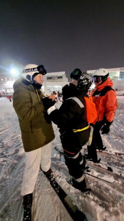 Three women in ski gear talking to each other on a ski hill at night.