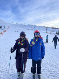 Two women at the base of a ski hill in ski gear wearing rainbow leis around their neck.