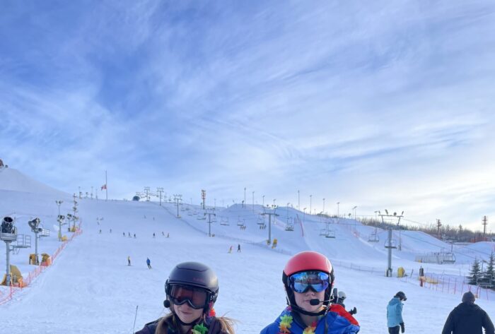 Two women at the base of a ski hill in ski gear wearing rainbow leis around their neck.