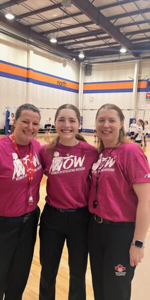 Three white women volleyball officials in pink shirts standing in front of a gym volleyball court.