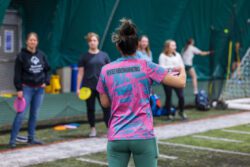 Women with her hair up in a bun and a pink and blue jersey showing a group of women how to hold a disc for disc golf.