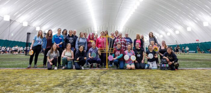 Group of women in athletic wear inside a fieldhouse dome taking a picture infront of a disc golf basket.
