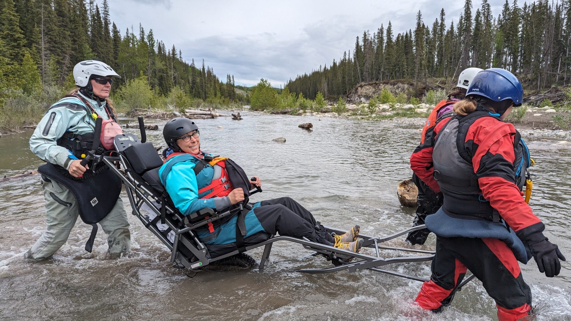 Four women crossing a river in wetsuits using an accessible transport carrier.