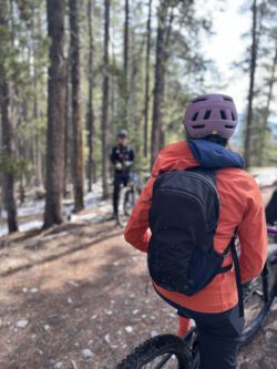 The back of a women with mountain biking gear listening to a blurred out coach in the forest.