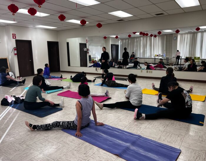 Group of women sitting on yoga mats in an indoor studio with mirrors stretching.