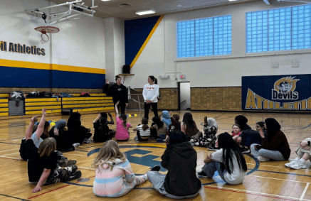 Group of girls sitting in a circle listening to an instructor in a gym.